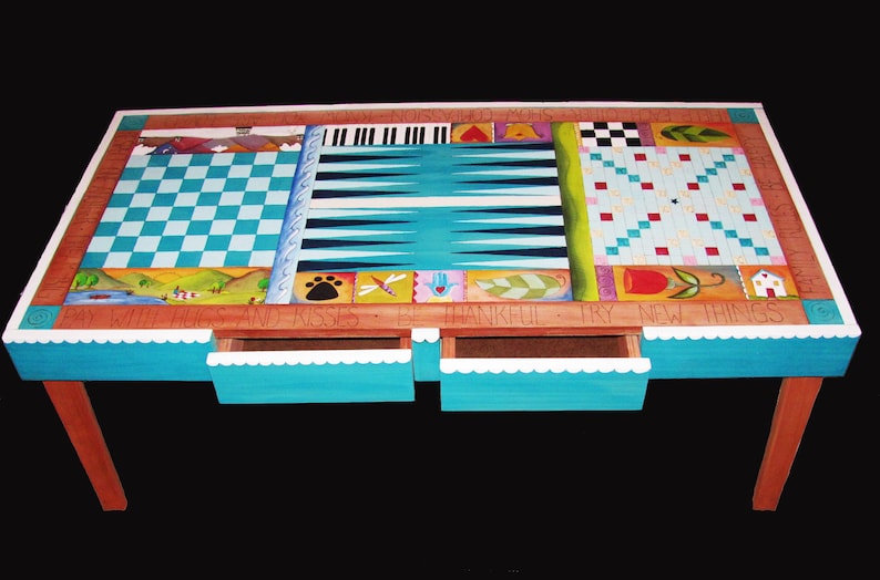 K&ouml;nnte beinhalten: Ein h&ouml;lzerner Spieltisch mit einer t&uuml;rkisblauen Tischplatte und braunen Beinen. Die Tischplatte zeigt ein Schachbrett, ein Backgammon-Brett und ein Tic-Tac-Toe-Brett. Der Tisch hat zwei Schubladen mit wei&szlig;en, gewellten Kanten. Der Tisch ist mit bunten Designs und den Worten "By With Hugs and Kisses Be Thankful Try New Things" bemalt.