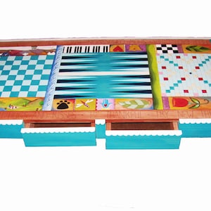 K&ouml;nnte beinhalten: Ein h&ouml;lzerner Spieltisch mit einem blau-wei&szlig;en Schachbrett, einem Backgammon-Brett und einem Tic-Tac-Toe-Brett. Der Tisch hat drei Schubladen mit einem wei&szlig;en, gewellten Rand und die Worte "By With Hugs And Kisses Be Thankful Try New Things" sind auf die Vorderseite gemalt.