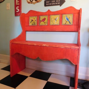May include: A bright orange wooden bench with a high backrest featuring four square tiles. Each tile displays a different bird illustration, including a goldfinch, a cardinal, a hummingbird, and a blue jay. The bench has a hinged seat lid.
