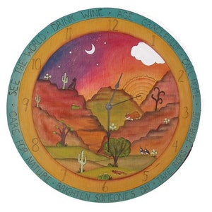 May include: A wooden clock with a colorful painted design of a desert landscape at sunset. The clock face has Roman numerals and the words "See the World, Drink Wine, Age Gracefully, Call Home, Care for Nature, Brighten Someone's Day, Take Risks, Forgive" around the edge.