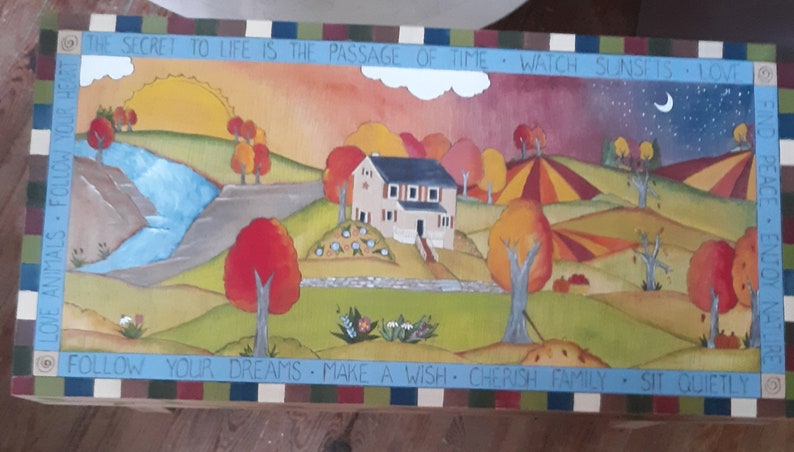 K&ouml;nnte beinhalten: Eine Holzkiste mit einem gemalten Bild eines Hauses in einer l&auml;ndlichen Landschaft. Die Kiste hat einen blau-gr&uuml;n-rot-wei&szlig; gestreiften Rand. Der Text auf der Kiste lautet: "Das Geheimnis des Lebens ist der Lauf der Zeit - Sonnenunterg&auml;nge beobachten - Tiere lieben - deinen Tr&auml;umen folgen - einen Wunsch machen - Familie sch&auml;tzen - ruhig sitzen - Frieden finden - die Natur genie&szlig;en."