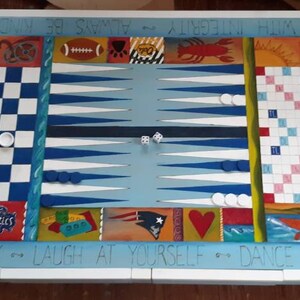 K&ouml;nnte beinhalten: Ein wei&szlig;er Holzspieltisch mit einem blau-wei&szlig;-roten Schachbrett, einem Backgammon-Brett und einem Scrabble-Brett. Der Tisch ist mit einem Design bemalt, das die Worte "Cherish Family - Laugh at Yourself - Dance and Sing" und "Integrity Always Be Kind Dream" enth&auml;lt.