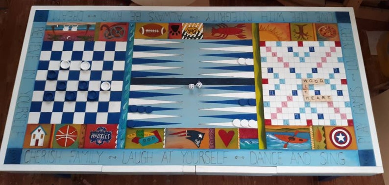 K&ouml;nnte beinhalten: Ein wei&szlig;er Holzspieltisch mit einem blau-wei&szlig;-roten Schachbrett, einem Backgammon-Brett und einem Scrabble-Brett. Der Tisch ist mit einem Design bemalt, das die Worte "Cherish Family - Laugh at Yourself - Dance and Sing" und "Integrity Always Be Kind Dream" enth&auml;lt.