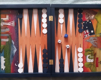 May include: A wooden backgammon game board with a blue frame. The board is open and shows the playing surface with black and white checkers. The board is decorated with painted scenes on each side, one with a cityscape and the other with a landscape.