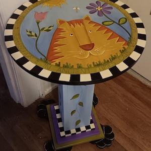 May include: A round wooden side table with a hand-painted cat face on the top, surrounded by a black and white checkered border. The text reads: IN ANCIENT TIMES CATS WERE WORSHIPED AS GODS THEY HAVE NOT FORGOTTEN THIS PURFECTION. It has a blue base.