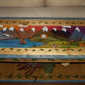 May include: A wooden table with a painted scene of mountains, a lake, and a house. The table has a blue and green border with a vine design. The text "SURFACE I THOUGHT IT MUST SURELY BE THE FAIREST PICTURE THE WHOLE EARTH AFFORDS" is painted on the table.