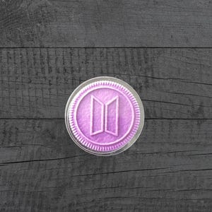 May include: A circular lavender-colored cookie-shaped object featuring a raised BTS logo in the center. The item has a textured surface with a ridged edge and is encased in a clear, thin plastic protective covering that follows its round shape.