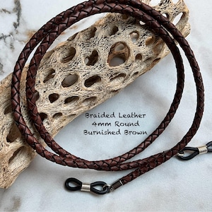 May include: Braided, burnished brown leather eyeglass chain. The 4mm round cord is designed to hold glasses. The chain is draped over a piece of driftwood. Text on the image reads "Braided Leather 4mm Round Burnished Brown."