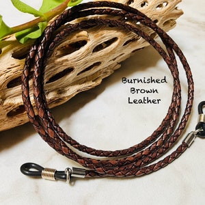 May include: A brown leather eyeglass chain with metal clasps. The chain is braided and has a burnished finish. "Burnished Brown Leather" is written on the chain.