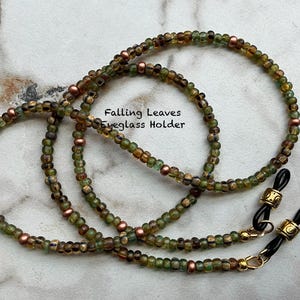 May include: An eyeglass holder chain featuring small, round, translucent green and brown beads interspersed with metallic copper-colored beads. The ends have gold-toned metal accents and black rubber loops. The text Falling Leaves Eyeglass Holder is visible.