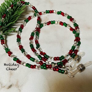 May include: A beaded eyeglass chain with red, green, and clear beads. The chain is designed with a holiday theme, and the text "Holiday Cheer" is visible. The chain has silver-colored attachments for glasses.