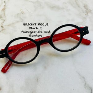 May include: Black and pomegranate red reading glasses with round frames. The glasses have a black frame front and red arms. The text "BRIGHT FOCUS Black & Pomegranate Red Readers" is visible.