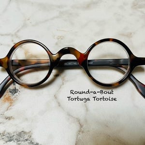 May include: Round eyeglasses with a tortoise shell pattern. The frames are round with a keyhole bridge. The temples are black and brown. The text "Round-a-Bout Tortuga Tortoise" is below the glasses.