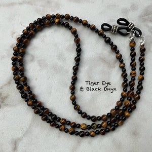 May include: Eyeglass chain made of tiger eye and black onyx beads. The chain has a mix of brown and black beads and includes the text "Tiger Eye & Black Onyx". The chain has silver-colored attachments for eyeglasses.