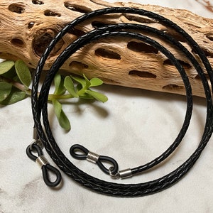 May include: Black leather braided glasses chain with silver metal clasps. The chain is lying on a wooden surface.