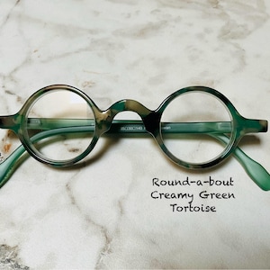May include: Round, creamy green tortoise eyeglasses with clear lenses. The glasses have a unique bridge design and slender arms. The text "Round-a-bout Creamy Green Tortoise" is written below the glasses.