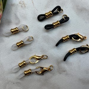 May include: Eyeglass holders in black and clear with gold-colored accents. The eyeglass holders have a loop on one end and a clasp on the other. The holders are designed to keep glasses secure and accessible.