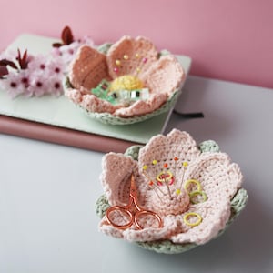 Może przedstawiać: Dwie ręcznie robione miski w kształcie kwiatów z szydełka w kolorze miękkiego różu i zieleni. Jedna miska zawiera akcesoria do szycia, takie jak szpilki i nożyczki. Druga miska zawiera małą szpulkę nici i inne materiały do szycia. Miski znajdują się na białej powierzchni.