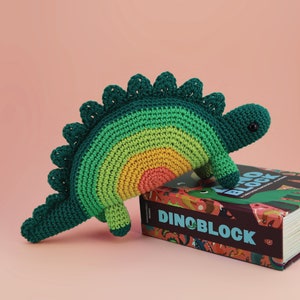 May include: A green, yellow, orange, and pink crocheted dinosaur toy is perched on top of a book titled "Dinoblock".