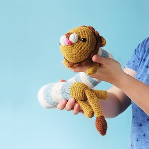 May include: A crocheted lion toy with a brown tail and blue and white striped body. The toy is being held by a person's hands.
