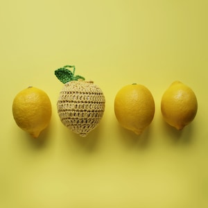 Puede incluir: Cuatro limones, uno de ellos tejido a crochet, sobre un fondo amarillo. El limón tejido a crochet está en el centro, con tres limones reales a cada lado.