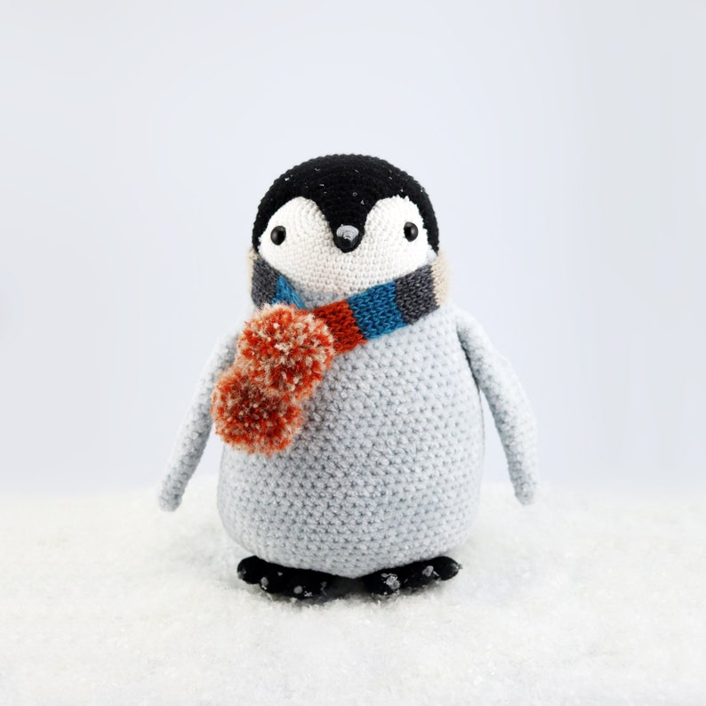 Puede incluir: Un juguete de ping&uuml;ino de crochet que lleva una bufanda a rayas azul, naranja y gris con un pomp&oacute;n naranja esponjoso. El ping&uuml;ino est&aacute; de pie sobre una superficie blanca.