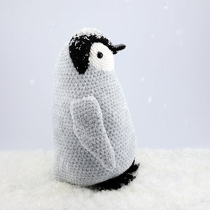 Puede incluir: Un juguete de ping&uuml;ino de crochet con una cara en blanco y negro y un cuerpo gris. El ping&uuml;ino est&aacute; de pie sobre una superficie blanca que parece nieve.