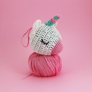Puede incluir: Una cabeza de unicornio de ganchillo blanco con una nariz rosa y un cuerno a rayas, descansando sobre una bola de hilo rosa. El unicornio tiene ojos cosidos negros y un lazo rosa para colgar. El fondo es rosa liso.