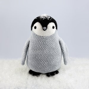 Puede incluir: Un peluche de ping&uuml;ino de crochet gris con ojos negros y un pico negro. El ping&uuml;ino est&aacute; sentado en una superficie blanca que parece nieve.