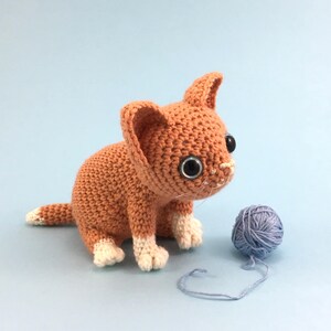 May include: A crocheted orange tabby kitten toy with white paws and black eyes. The kitten is sitting on a light blue background and is looking to the right. A ball of blue yarn is in the foreground.