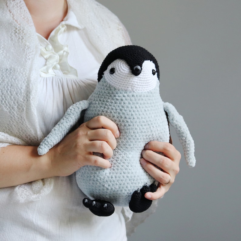 Puede incluir: Un juguete de peluche de ping&uuml;ino de crochet con un cuerpo azul claro, patas negras y una cara blanca y negra. El ping&uuml;ino est&aacute; siendo sostenido por una persona.