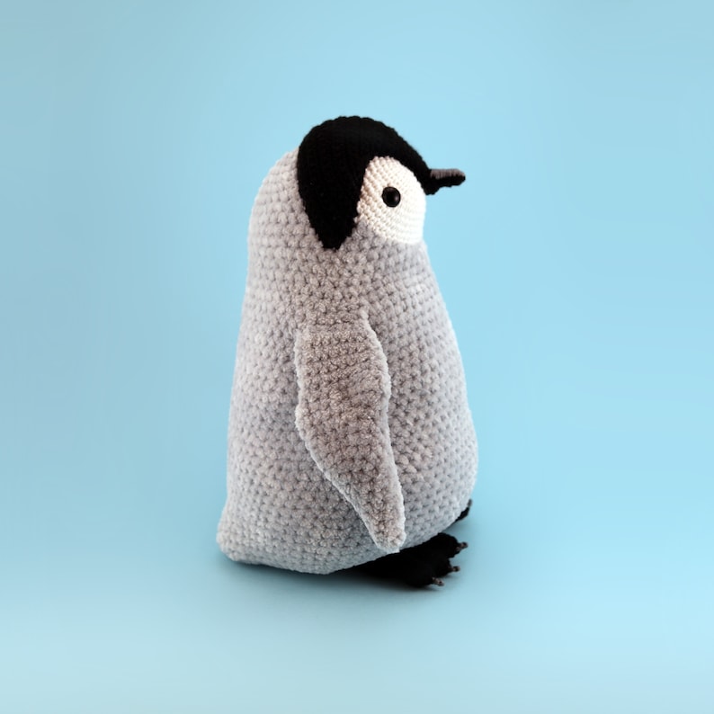 Puede incluir: Un juguete de peluche de ping&uuml;ino de crochet gris con ojos negros y un pico negro. El ping&uuml;ino est&aacute; de pie sobre un fondo azul claro.