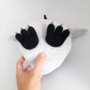 Puede incluir: Un animal de peluche blanco tejido a crochet con pies negros tejidos a crochet. El animal est&aacute; acostado de espaldas con los pies en el aire.
