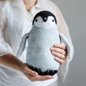 Puede incluir: Un juguete de peluche de ping&uuml;ino de crochet con un cuerpo azul claro, ojos negros y un pico negro. El ping&uuml;ino est&aacute; siendo sostenido por las manos de una persona.