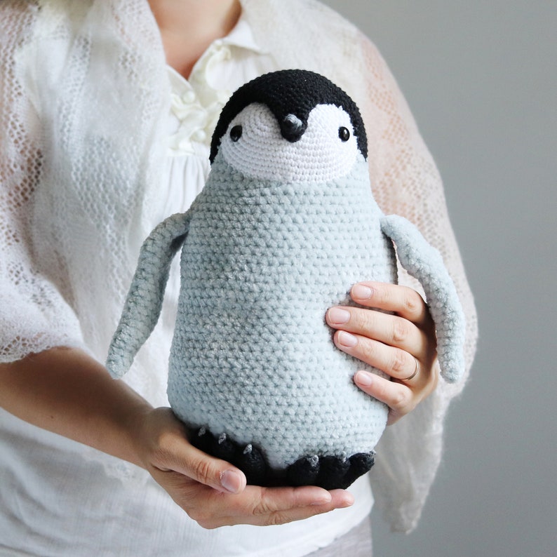 Puede incluir: Un juguete de peluche de ping&uuml;ino de crochet con un cuerpo azul claro, ojos negros y un pico negro. El ping&uuml;ino est&aacute; siendo sostenido por las manos de una persona.