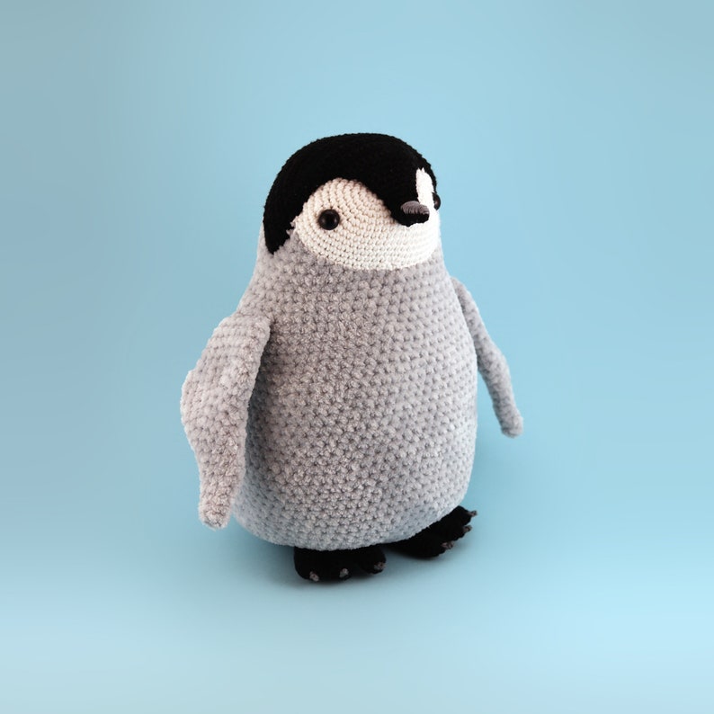 Puede incluir: Un juguete de ping&uuml;ino de crochet con un cuerpo gris, una cabeza negra y una cara blanca. El ping&uuml;ino tiene ojos negros y un pico negro.