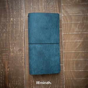 May include: A teal leather journal with a textured finish, lying open on a wooden surface. The journal is secured with a black elastic band. The brand name "mindh." is printed in white at the bottom.