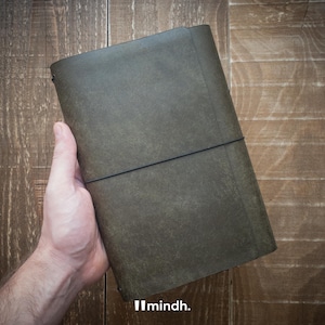 Könnte beinhalten: Ein dunkel olivgrünes Lederjournal mit einem schwarzen Gummibandverschluss. Das Journal wird in einer Hand gehalten und zeigt seine strukturierte Oberfläche. Der Markenname "mindh." ist unten rechts zu sehen.