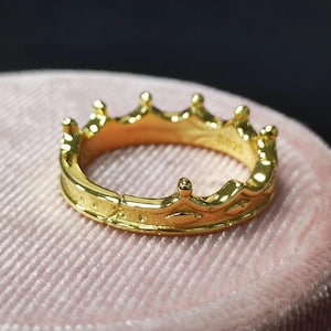 May include: A gold crown ring with a detailed design. The ring is on a pink velvet surface.
