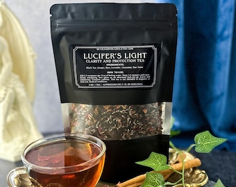 Pode incluir: Uma embalagem preta de "Lucifer's Light Clarity and Protection Tea" com uma janela transparente, uma chávena de chá cheia, paus de canela e folhas decorativas. O chá é de cor âmbar escuro. A embalagem tem um rótulo com texto.