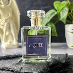 May include: A clear glass perfume bottle with a gold cap and a faceted crystal top. The label reads "LUCIFER" in gold script on a navy blue background. The bottle is 1.7 fl oz (50 ml).