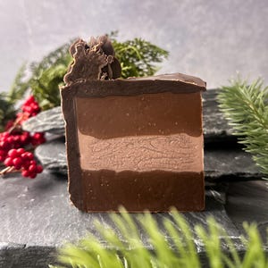 May include: A layered bar of soap with shades of brown, resembling a chocolate dessert. The soap has a dark chocolate top with a textured design and a lighter brown middle layer. The soap is displayed on a dark gray surface with greenery and red berries.