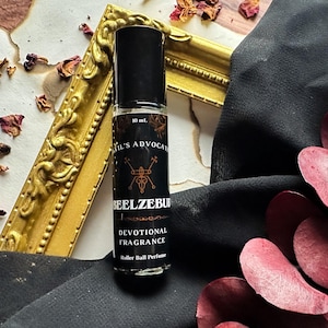 May include: A black rollerball perfume bottle with a black and white label that reads "Beelzebub Devotional Fragrance" and "Devil's Advocate". The bottle is sitting on a wooden surface with red and brown dried flowers.