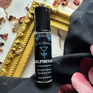 May include: A black rollerball perfume bottle with a blue and white label. The label reads "Evil's Advocate", "Belphegor", "Devotional Fragrance", and "Roller Ball Perfume". The bottle is sitting on a gold frame and a black fabric surface.
