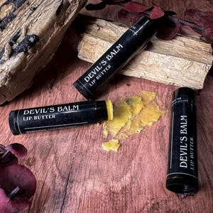 May include: Two black lip balm tubes with the text "Devil's Balm Lip Butter" on a wooden surface. One tube is open and the lip balm is partially used.