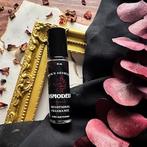 Puede incluir: Un frasco de perfume negro de bola con una etiqueta roja y negra. La etiqueta dice "Asmodeus Devotional Fragrance" y "Devil's Advocate" con un sello. El frasco es de 10 ml.