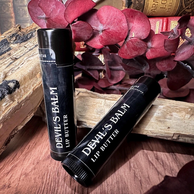 May include: Two black lip balm tubes with white text that reads "Devil's Balm Lip Butter". The tubes are on a wooden surface with red leaves in the background.