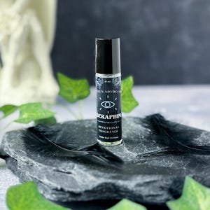 May include: A black rollerball perfume bottle with the text "SERAPHIM DEVOTIONAL FRAGRANCE" and an eye symbol. The bottle is on a dark grey stone with black feathers.