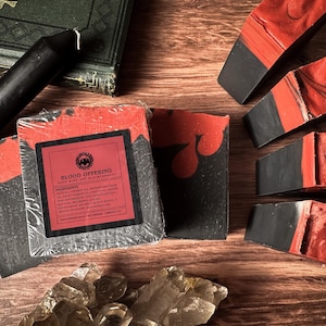 Puede incluir: Barras de jabón artesanales con un llamativo diseño rojo y negro. Las barras están etiquetadas como "Blood Offering" y están dispuestas sobre una superficie de madera con una vela negra, un libro y cristales. El jabón tiene un aspecto veteado.