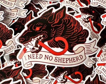 May include: A black and red wolf head sticker with a red curved line coming out of its mouth. The sticker has a white banner with the text "I NEED NO SHEPHERD".